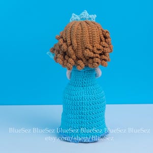 May include: A handmade crochet doll, viewed from the back, featuring a light blue dress and a brown curly hairstyle. The doll has a small blue crown. The background is a solid light blue. The doll is sitting on a white surface.