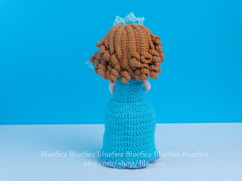 May include: A handmade crochet doll, viewed from the back, featuring a light blue dress and a brown curly hairstyle. The doll has a small blue crown. The background is a solid light blue. The doll is sitting on a white surface.