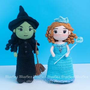 May include: Two handmade crochet dolls: a green-skinned witch in a black dress and hat, holding a broom, and a princess in a blue dress with a crown and wand. The dolls are set against a bright blue background.