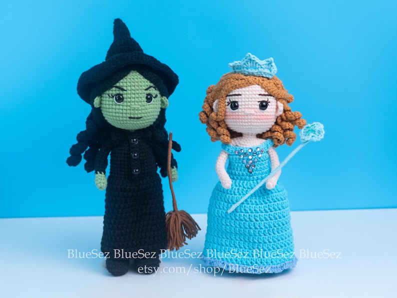 May include: Two handmade crochet dolls: a green-skinned witch in a black dress and hat, holding a broom, and a princess in a blue dress with a crown and wand. The dolls are set against a bright blue background.