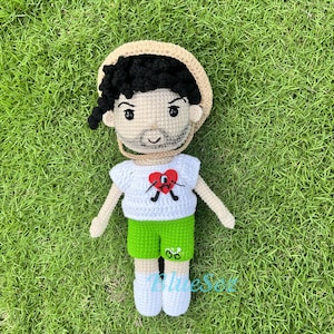 Puede incluir: Una mu&ntilde;eca de crochet con un sombrero de paja, una camisa blanca con un coraz&oacute;n rojo y pantalones cortos verdes. La mu&ntilde;eca tiene el pelo negro y una barba marr&oacute;n.