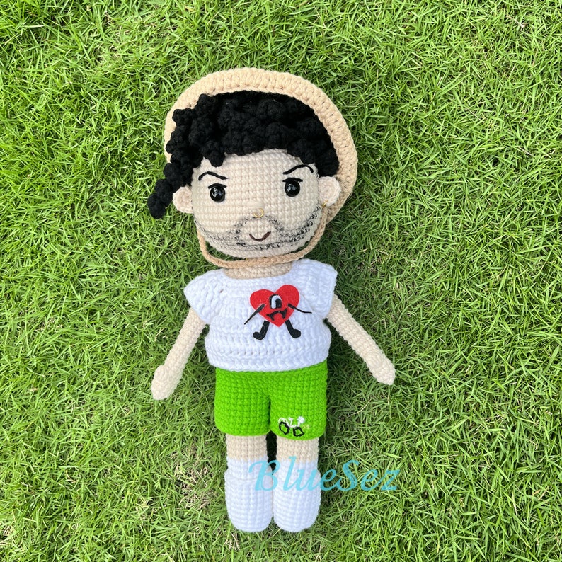 Puede incluir: Una mu&ntilde;eca de crochet con un sombrero de paja, una camisa blanca con un coraz&oacute;n rojo y pantalones cortos verdes. La mu&ntilde;eca tiene el pelo negro y una barba marr&oacute;n.