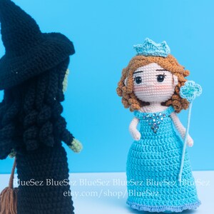May include: Two crocheted dolls: a black witch with a pointed hat and a blue princess with a crown. The witch has a broom, and the princess holds a wand. The dolls are set against a solid blue background.