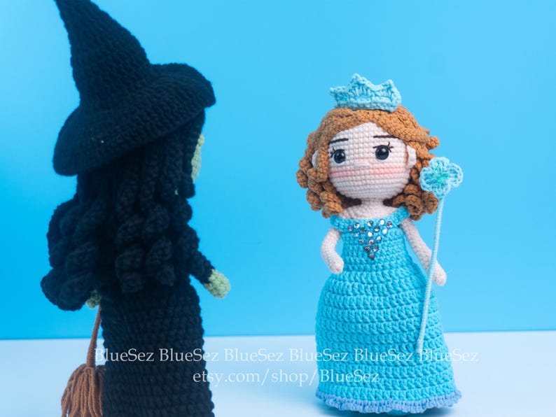 May include: Two crocheted dolls: a black witch with a pointed hat and a blue princess with a crown. The witch has a broom, and the princess holds a wand. The dolls are set against a solid blue background.