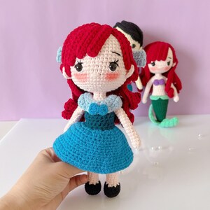 May include: Crocheted doll with red hair, blue dress, and black shoes. The doll has a light pink face with black eyes and eyelashes. Two other dolls are in the background, one with black hair and another with red hair and a mermaid tail. The dolls are handmade.