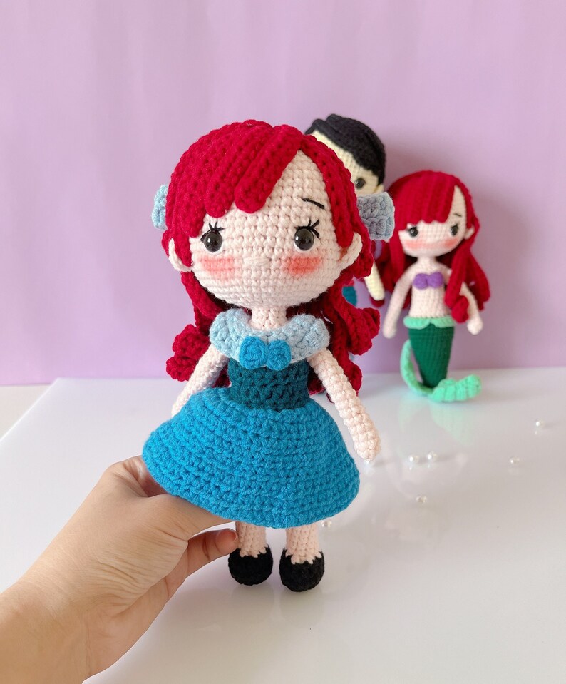 May include: Crocheted doll with red hair, blue dress, and black shoes. The doll has a light pink face with black eyes and eyelashes. Two other dolls are in the background, one with black hair and another with red hair and a mermaid tail. The dolls are handmade.