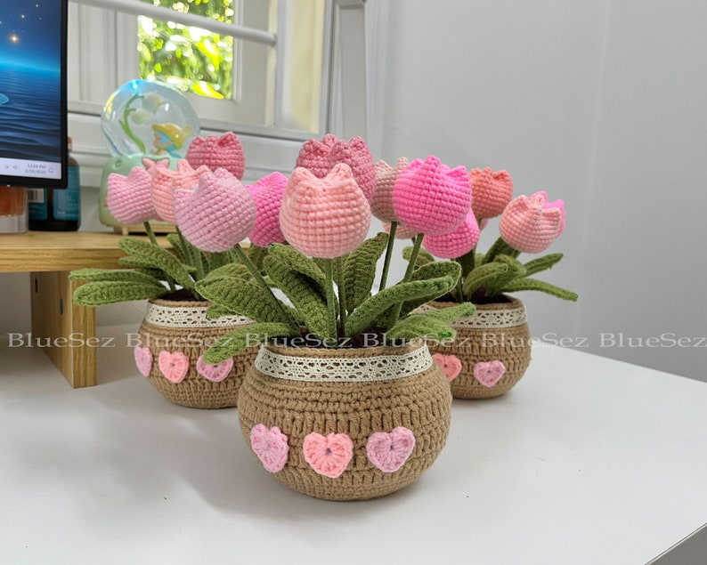 May include: Three crocheted flower pots with pink flowers and hearts. The pots are made of brown yarn and have a white lace trim.