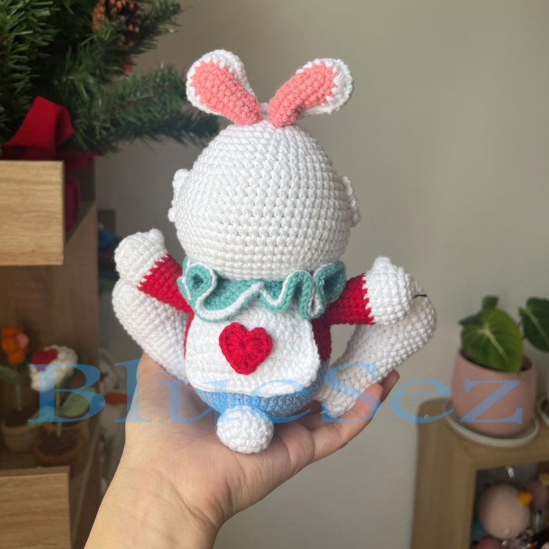 May include: A white crocheted bunny rabbit wearing a red and green outfit with a heart-shaped bib. The bunny has pink ears and is holding its arms up.