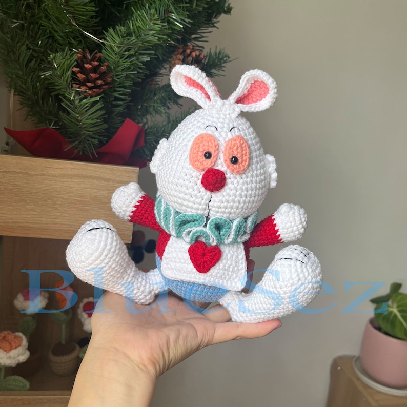 May include: White crocheted rabbit plush toy with red, green, and blue accents. The rabbit has a heart-shaped patch on its chest and a green and white scarf around its neck.