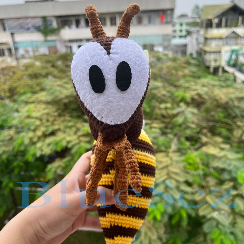 Puede incluir: Un juguete de abeja de crochet con rayas marrones y amarillas, una cabeza marr&oacute;n y ojos de fieltro blanco. La abeja tiene una cara en forma de coraz&oacute;n.
