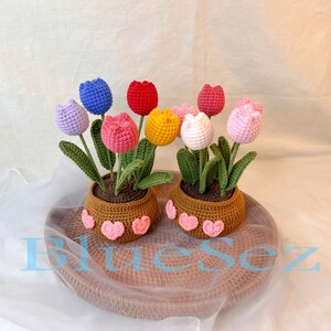 May include: Two crocheted flower pots with colorful crocheted tulips. The pots are brown and have pink hearts around the top. The pots are sitting on a woven brown tray.