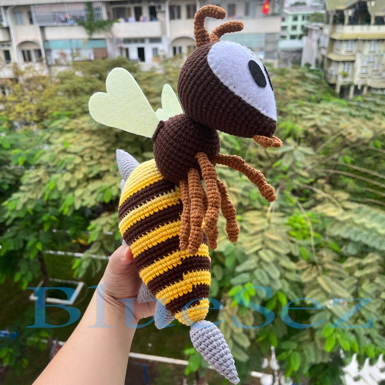 Puede incluir: Un juguete de abeja de crochet con rayas marrones y amarillas, patas marrones y un aguij&oacute;n gris. La abeja tiene una cabeza marr&oacute;n con una cara blanca y ojos negros. La abeja tiene dos alas de color amarillo claro.