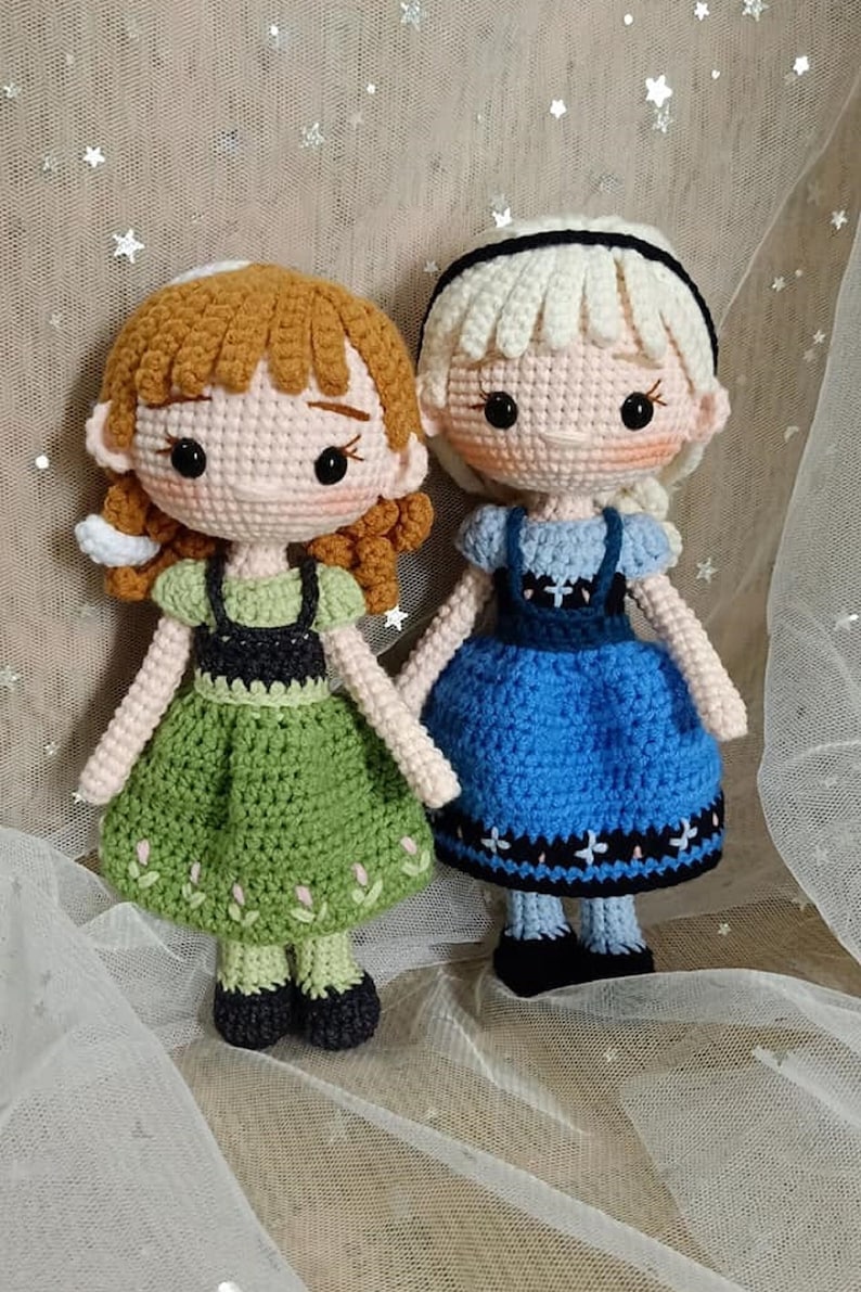 May include: Two crocheted dolls dressed as the characters Anna and Elsa from the movie Frozen. Anna is wearing a green dress with a black bodice and brown hair. Elsa is wearing a blue dress with a black bodice and blonde hair.
