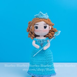 May include: Crocheted doll of a princess in a blue dress, crown, and wand. The doll has brown hair and rosy cheeks. The background is a solid blue. The doll is handmade and has a unique design.