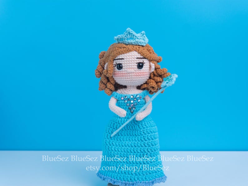 May include: Crocheted doll of a princess in a blue dress, crown, and wand. The doll has brown hair and rosy cheeks. The background is a solid blue. The doll is handmade and has a unique design.