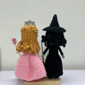 May include: Crocheted dolls of a princess and a witch. The princess wears a pink dress and a silver crown. The witch is dressed in black with a pointed hat. Both dolls are standing on a wooden base.