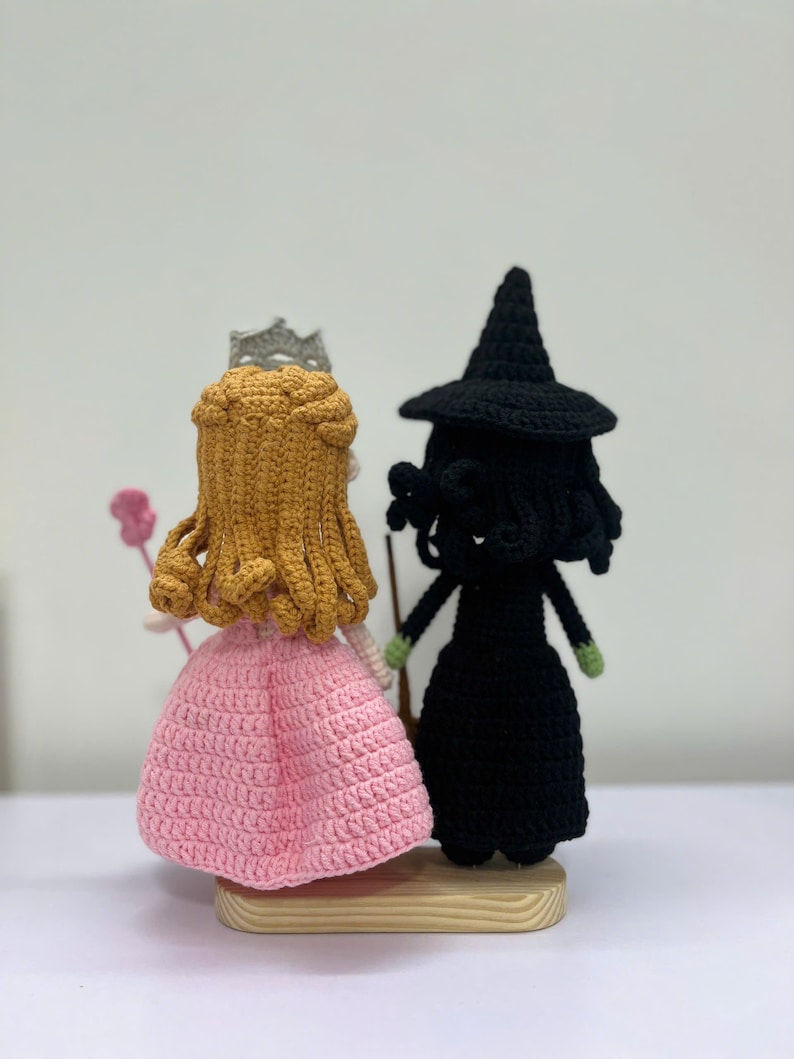 May include: Crocheted dolls of a princess and a witch. The princess wears a pink dress and a silver crown. The witch is dressed in black with a pointed hat. Both dolls are standing on a wooden base.