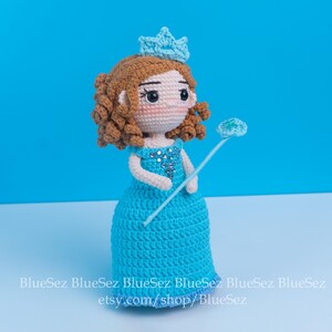 May include: A handmade crochet doll of a princess, featuring a blue dress, a crown, and a wand. The doll has brown curly hair and is adorned with silver embellishments. The background is a solid light blue.