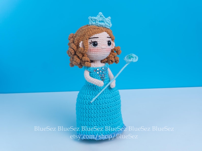 May include: A handmade crochet doll of a princess, featuring a blue dress, a crown, and a wand. The doll has brown curly hair and is adorned with silver embellishments. The background is a solid light blue.