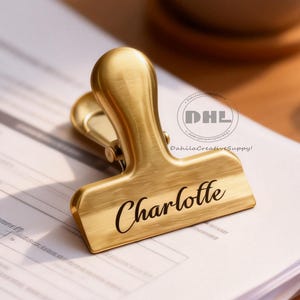 May include: A gold-coloured paper clip, engraved with the name "Charlotte", sits atop a stack of white papers. A cup of coffee and a laptop are visible in the background, suggesting an office or workspace.