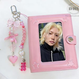 Pode incluir: Um álbum de fotos rosa com uma foto de uma pessoa com cabelo loiro, um chaveiro com contas rosa, um coração rosa e um pingente de ursinho de goma. O álbum tem recortes em forma de estrela e um fecho de pressão prateado. Decorações douradas em forma de estrela e laços rosa estão espalhados.