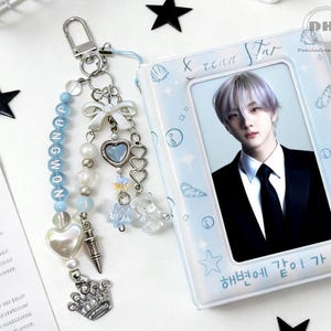 May include: A light blue photo album featuring a person in a black suit and tie. A keychain with blue beads, charms, and a crown charm is next to the album. The album has text in Korean. Black star-shaped decorations are scattered around.
