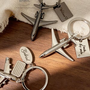 May include: Silver airplane keychains with personalized tags. One keychain has the name "William" engraved, another says "Jack, Fly safe", and the third has the name "Gabriel". The keychains are on a wooden surface.