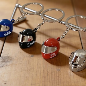 Puede incluir: Cuatro llaveros de mini cascos de carreras en azul, negro, rojo y plateado. Cada casco tiene una visera plateada y está unido a un llavero. El casco azul tiene "M&B", el negro "Ethan", el rojo "Jerry 11.12.23" y el plateado "Will" grabados.