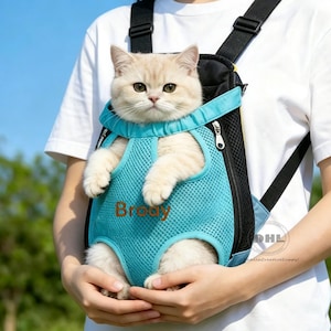 Puede incluir: Una mochila transportadora de malla azul claro para mascotas con correas negras ajustables y paneles laterales negros. El transportador tiene una abertura frontal para que un gato se siente con las patas delanteras hacia afuera. El nombre Brody está bordado en marrón.