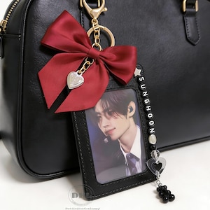 Könnte beinhalten: Schwarze Handtasche mit einem roten Satin-Schleifen-Schlüsselanhänger. Der Schlüsselanhänger hat einen Herzanhänger und einen Fotokartenhalter mit dem Namen "SUNGHOON" in Perlen geschrieben. Der Fotokartenhalter hat ein Foto einer Person.