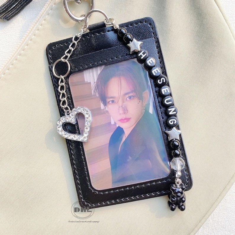 May include: Black leather ID card holder with a photo of a person. The holder has a silver chain with a rhinestone heart charm and a beaded chain with the name "HEESEUNG" and a black bear charm.