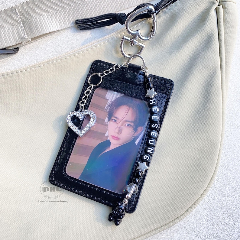 May include: Black cardholder with a photo of a person, a silver heart charm, and a beaded chain with the name "HEESEUNG". The cardholder is attached to a silver heart-shaped clasp.