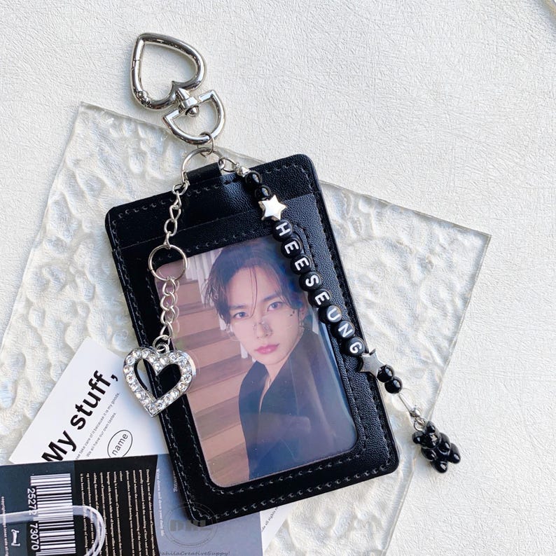 May include: Black leather ID card holder with a photo of a person. The holder has a silver heart-shaped keychain and a beaded charm that spells out "HEESEUNG". A small black bear charm hangs from the holder.