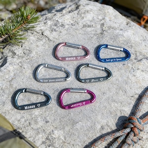 May include: Six colorful carabiner clips with engraved text and heart symbols. The colors include pink, blue, green, and purple. The text includes names and phrases like "Let's go to Space". The clips are on a gray rock surface.