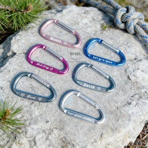 May include: A collection of colorful carabiner clips in pink, purple, blue, and silver. Each clip has a silver gate and is engraved with text, including names and phrases like "Let's go to space" and "Your phone number". The clips are displayed on a rock surface.