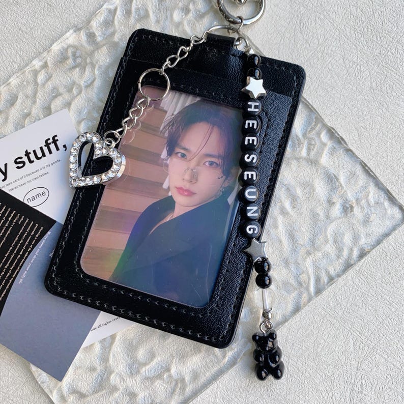 May include: Black leather ID card holder with a photo of a person. The holder has a silver chain with a rhinestone heart charm and a beaded charm with the name "HEESEUNG" and a black gummy bear. A silver keyring is attached.