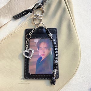 May include: Black leather ID holder with a photo of a person. The holder has a silver chain with a rhinestone heart charm and a beaded strap with the name "HEESEUNG". A black bear charm hangs from the bottom. The holder is attached to a beige bag.