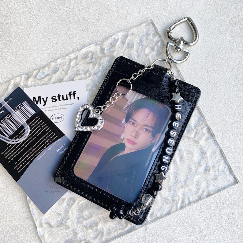 May include: Black ID card holder with a photo of a person, a silver heart charm, and a beaded chain. The holder has the name "HEESEUNG" spelled out in white beads. A small card with the text "My stuff" is also visible.