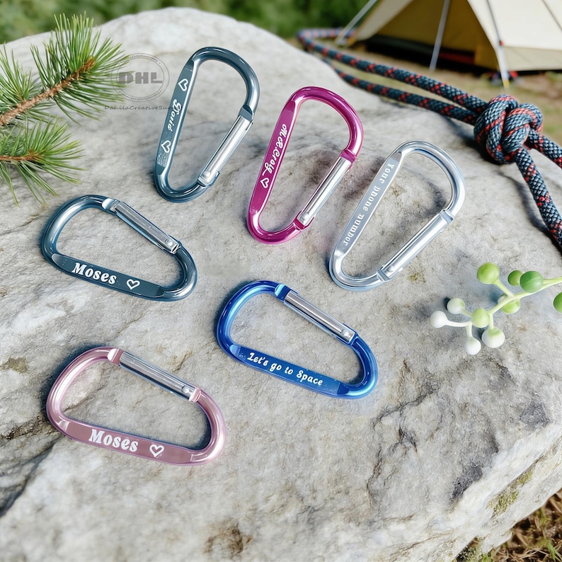 May include: An assortment of colorful carabiner clips in various shades of blue, pink, and silver, resting on a gray stone surface. Each clip has a personalized inscription, such as names and phrases like "Let's go to Space".