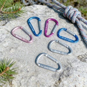 May include: A collection of colorful carabiners in pink, blue, and purple, with silver accents. Each carabiner has a unique inscription, including names and phrases like "Let's go to space" and "Your phone number". The carabiners are arranged on a textured stone surface.