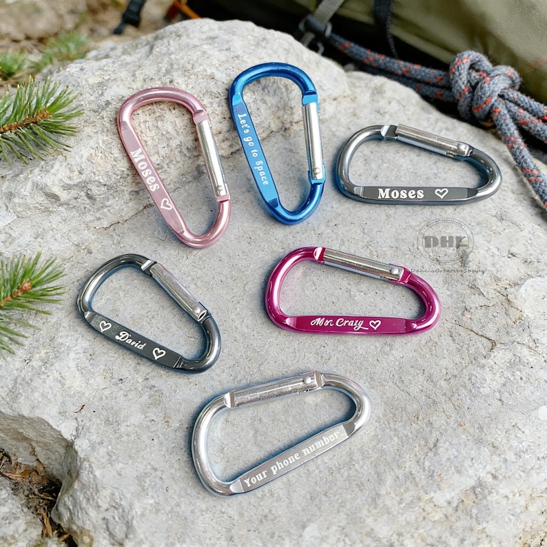 May include: A collection of six carabiner clips in various colors, including pink, blue, gray, and magenta. Each clip has a silver gate and personalized text, such as names and phrases like "Let's go to Space" and "Your phone number."