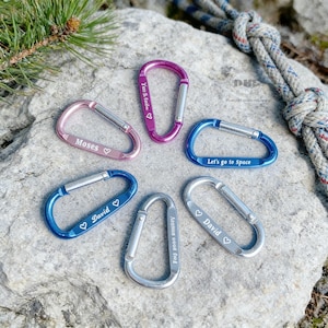 May include: A collection of colorful carabiner clips in pink, purple, blue, and silver. Each clip has a silver gate and features engraved text, including names and phrases like "Let's go to Space". The clips are arranged on a gray rock surface.