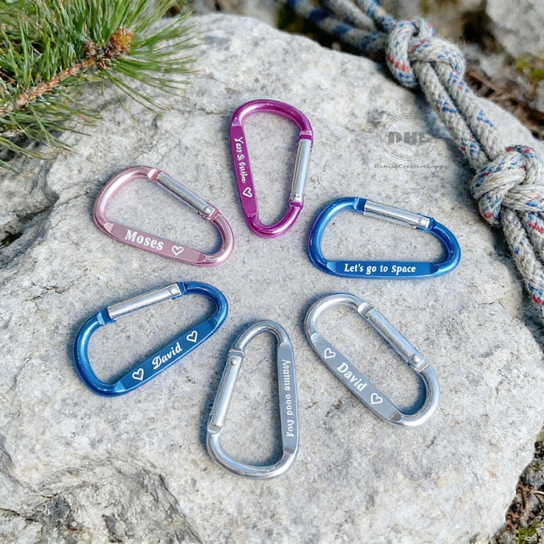 May include: A collection of colorful carabiner clips in pink, purple, blue, and silver. Each clip has a silver gate and features engraved text, including names and phrases like "Let's go to Space". The clips are arranged on a gray rock surface.