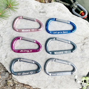 May include: Six colorful carabiner clips in pink, blue, purple, and silver. Each clip has a silver gate and is engraved with text, including names and phrases like "Let's go to Space" and "Your phone number". The clips are displayed on a stone surface.