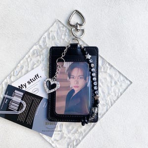 May include: Black ID card holder with a photo, a silver heart charm, and a beaded strap with the name "HEESEUNG". The holder has a heart-shaped clasp and a chain. A card with the text "My stuff" is visible.
