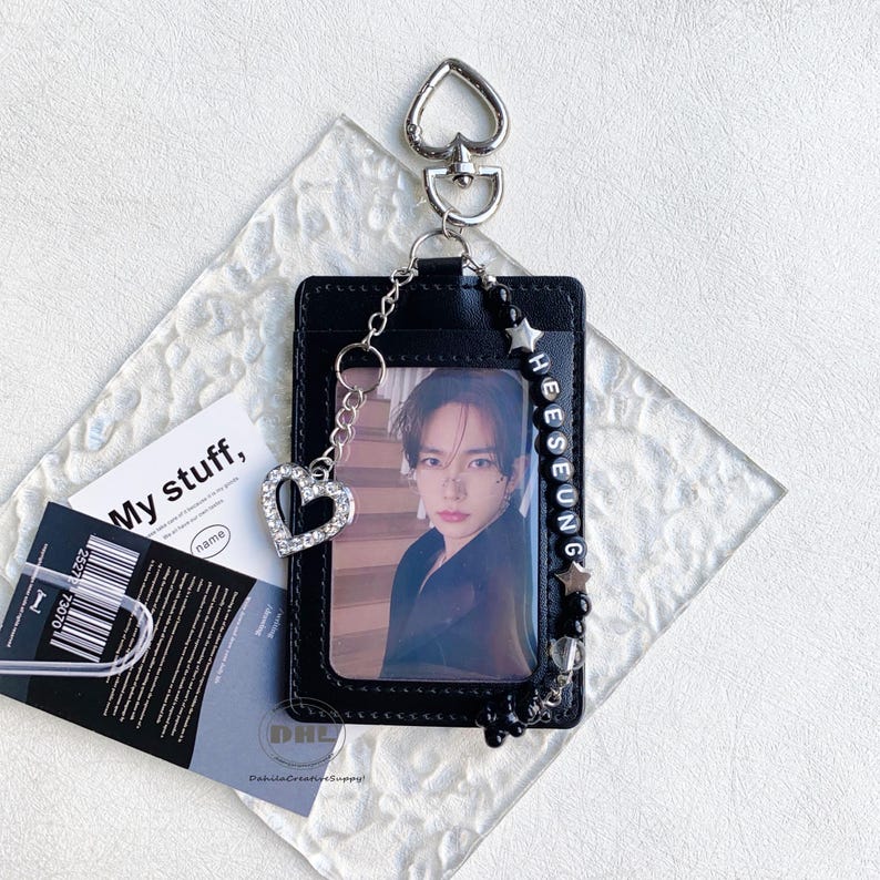 May include: Black ID card holder with a photo, a silver heart charm, and a beaded strap with the name "HEESEUNG". The holder has a heart-shaped clasp and a chain. A card with the text "My stuff" is visible.