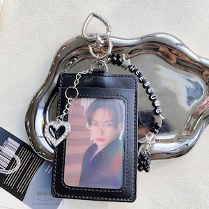 May include: Black leather ID card holder with a photo of a person. The holder has a silver chain with a rhinestone heart charm. A beaded charm with the name "HEESEUNG" is attached, along with a small black bear charm.