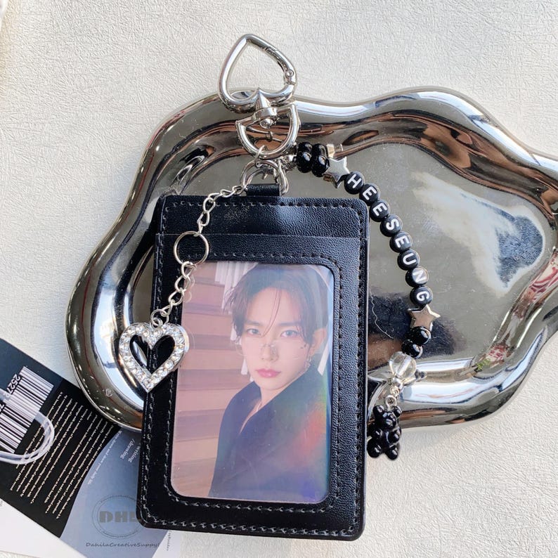 May include: Black leather ID card holder with a photo of a person. The holder has a silver chain with a rhinestone heart charm. A beaded charm with the name "HEESEUNG" is attached, along with a small black bear charm.