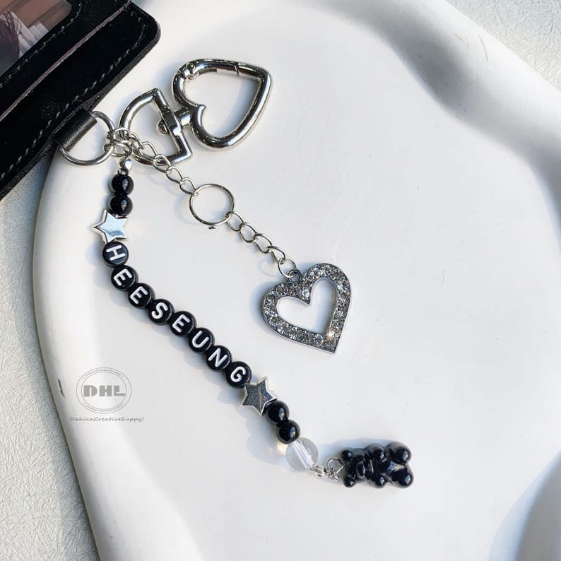 May include: A silver keychain with a heart-shaped clasp and a dangling charm. The charm features a rhinestone-studded heart, black beads spelling "HEESEUNG", star-shaped beads, and a black gummy bear charm. The keychain is on a white surface.