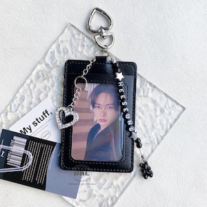 May include: Black cardholder with a photo, a silver heart-shaped charm, and a beaded charm with the name "HEESEUNG". The cardholder has a silver heart-shaped keychain. A small black bear charm hangs from the beaded charm.